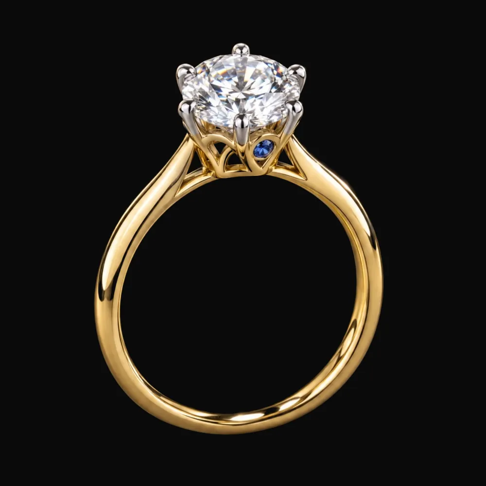 Le solitaire Comtesse de face sur fond noir : anneau en or jaune, diamant rond tenu par six griffes en or blanc, petit saphir bleu dans la corbeille